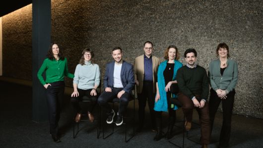 Die sieben Theatertreffen-Jurymitglieder stehen oder sitzen for einer Kieselwand und schauen in die Kamera.