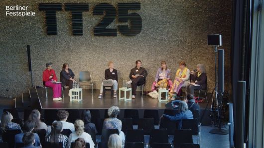 7 Menschen sitzen auf der Bühne, eine Person spricht und die übrigen schauen sie an.