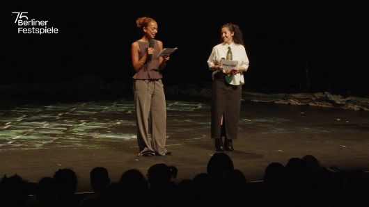 Zwei Personen stehen auf einer Bühne der Berliner Festspiele und halten Moderationskarten in den Händen. Im Vordergrund ist das Publikum sichtbar.