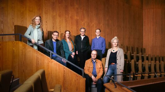Die sieben Theatertreffen-Jurymitglieder stehen oder sitzen im Rang eines Theaters und schauen in die Kamera.
