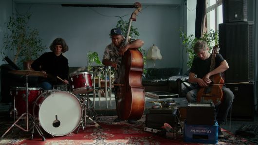 Drei Musiker*innen mit Schlagzeug, Kontrabass und Cello in einem Raum mit Pflanzen, Teppich und Lautsprechern