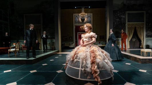Eine junge Frau in einem Reifrock steht in einem Salon, im Hintergrund sind weitere Personen in historischen Kostümen