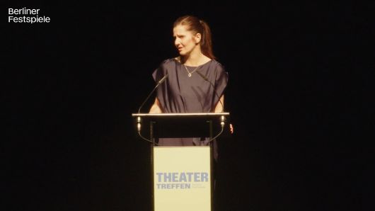 Nora Hertlein-Hull standing at a lectern, which has Theatertreffen written on it