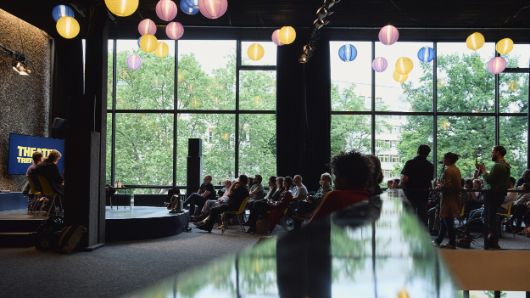 Seitlicher Blick auf das Publikumsgespräch im  Oberen Foyer. Links die Bühne, in der Mitte und rechts das Publikum, im Vordergrund unscharf ein Geländer, in dem sich die Lampions an der Decke und die Fester im Hintergrund spiegeln.
