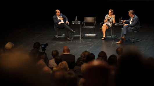 Drei Personen sitzen auf einer Bühne und diskutieren vor Publikum.
