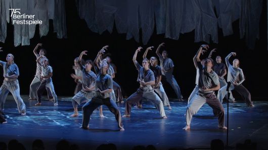 Ensemble von Tanzenden in grauen und weißen Kostümen auf einer Bühne. Die Gruppe führt eine synchronisierte Choreografie mit ausdrucksstarken Armbewegungen unter blauem Licht aus. Über der Bühne hängen stoffartige Elemente. 