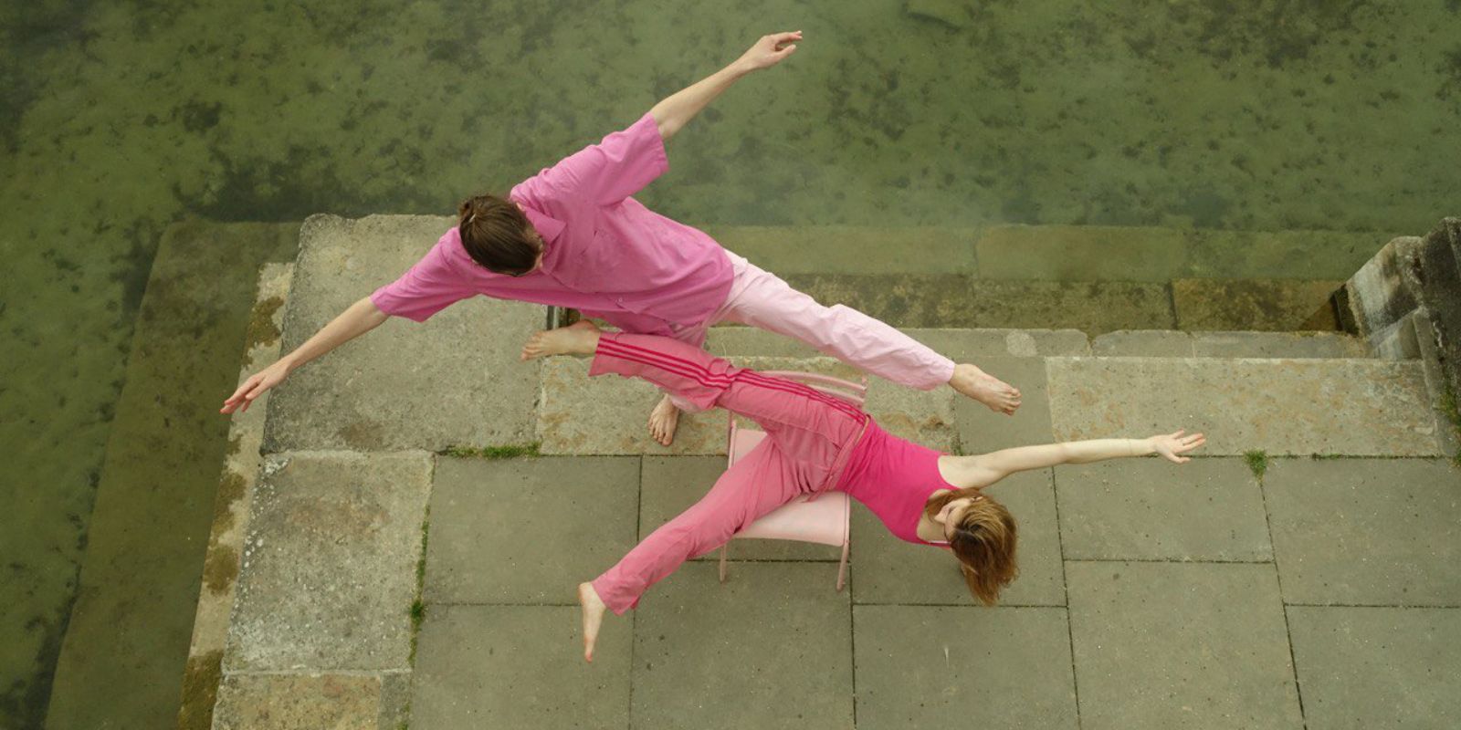 Zwei Performer*innen von oben fotografiert. Sie tragen pinke Kleidung und machen große Bewegungen auf einem Stuhl.