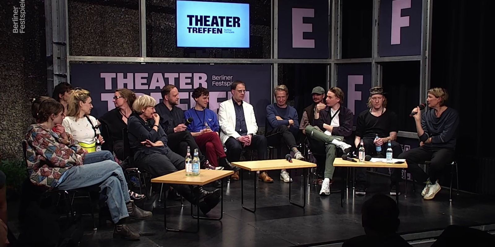 Eine Gruppe von Personen sitz um drei kleine Tische herum. Hinter ihnen eine Gerüst mit dem Logo des Theatertreffen.