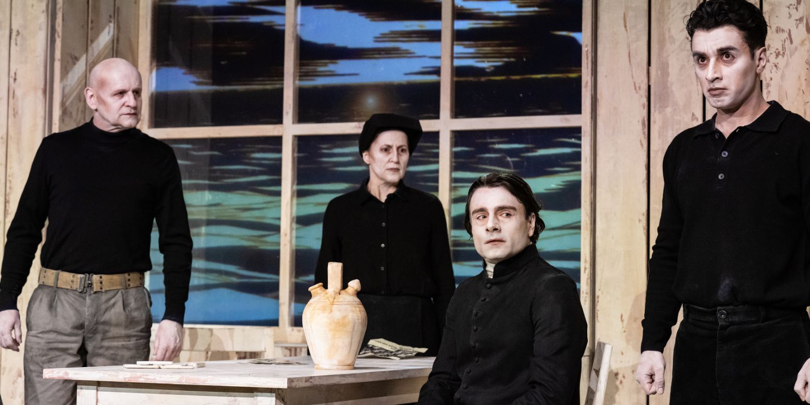 Four people in old-fashioned-looking black clothes are standing in a kitchen table scene where everything is made of wood.