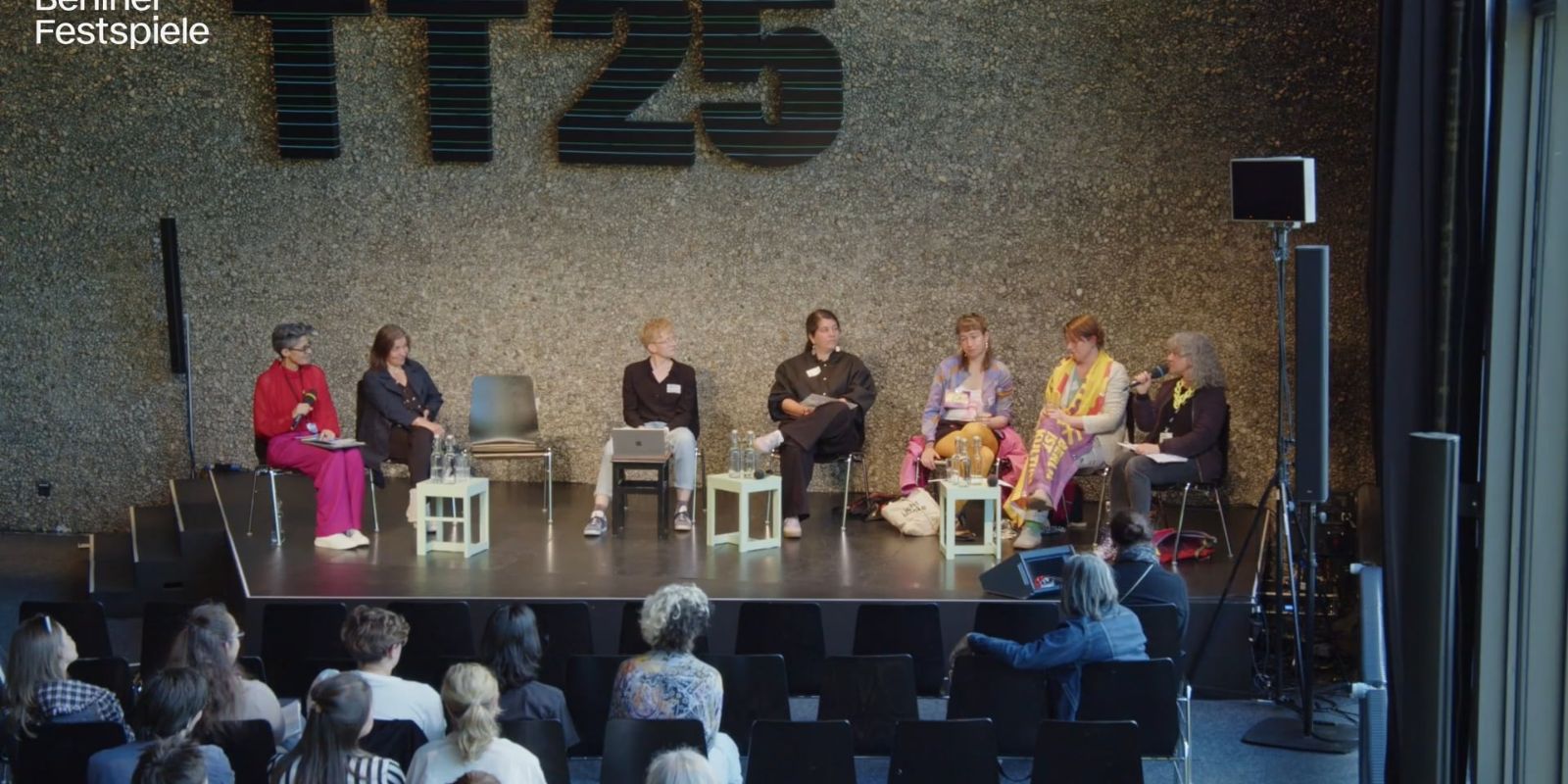 7 Menschen sitzen auf der Bühne, eine Person spricht und die übrigen schauen sie an.