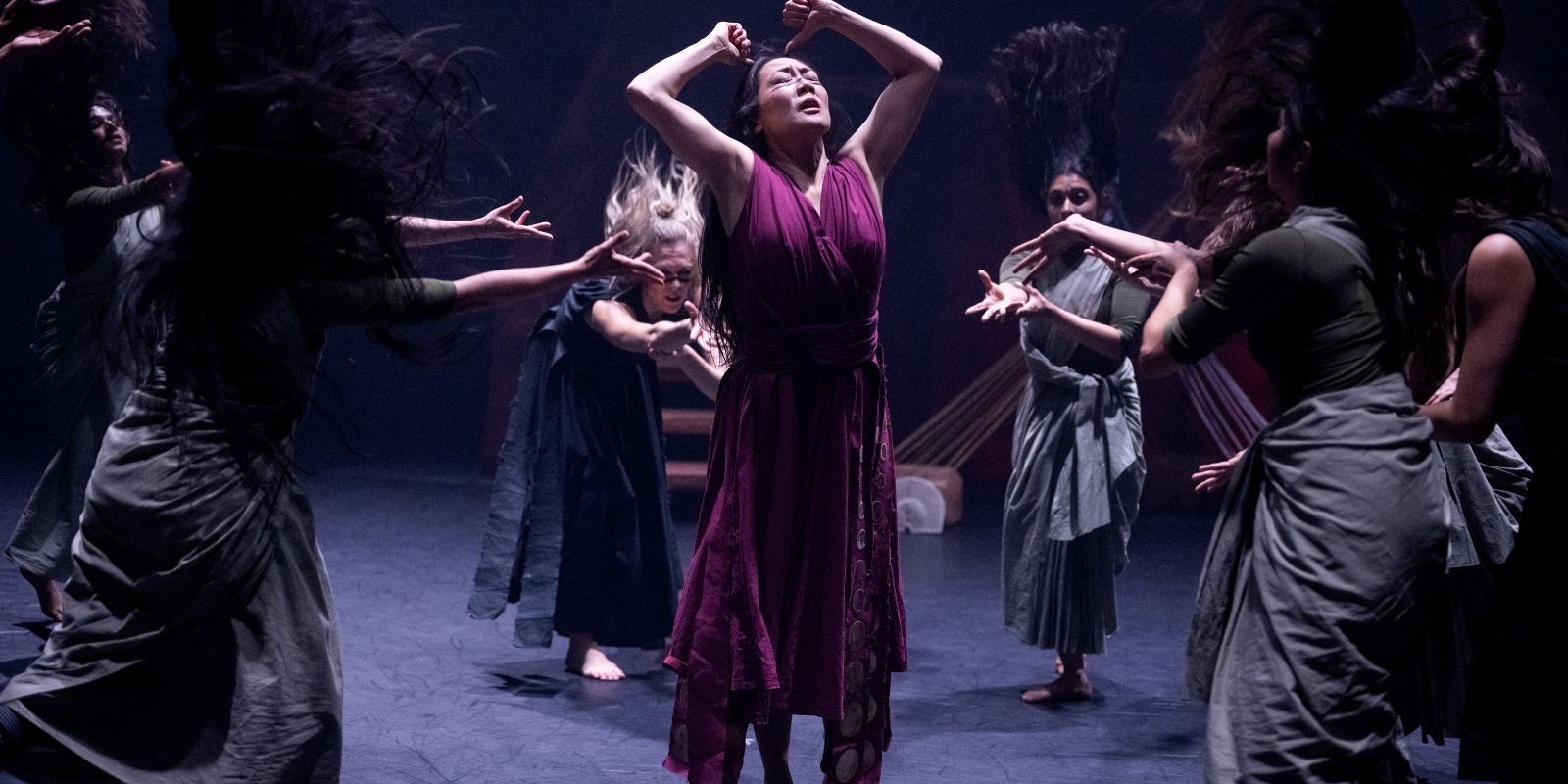 A dancer stands in the middle of a group of other dancers. She raises her fists and turns her face towards the sky.