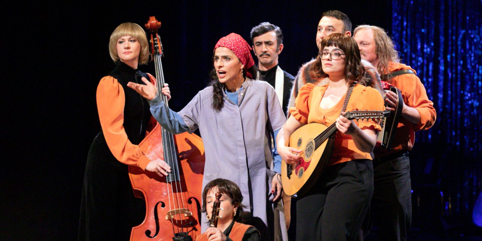 A woman wearing a headscarf and a work coat stands in the middle of a group of musicians and speaks to the audience.