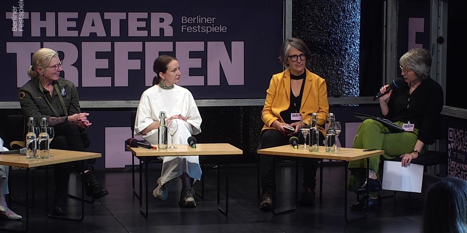 Vier Frauen sitzen an zwei kleinen Tischen, hinter ihnen der Schriftzug Theatertreffen. Die Frau ganz rechts hat ein Mikrofon in der Hand.