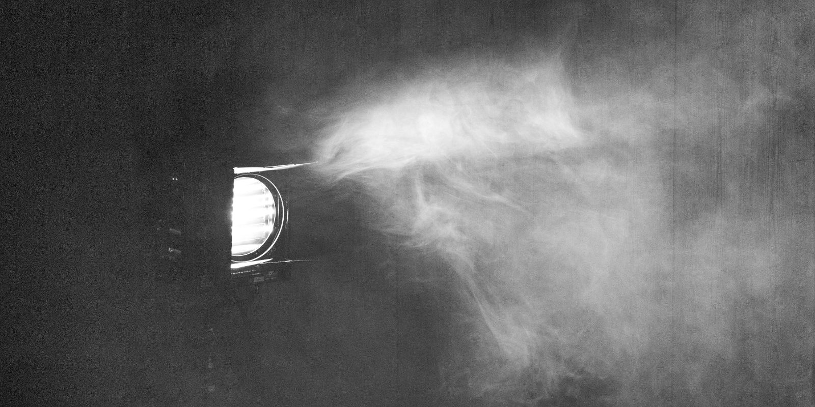 Schwarz-weißes Bild eines Scheinwerfers, der nach links strahlt, Nebel oder Rauch im Lichtkegel sichtbar.