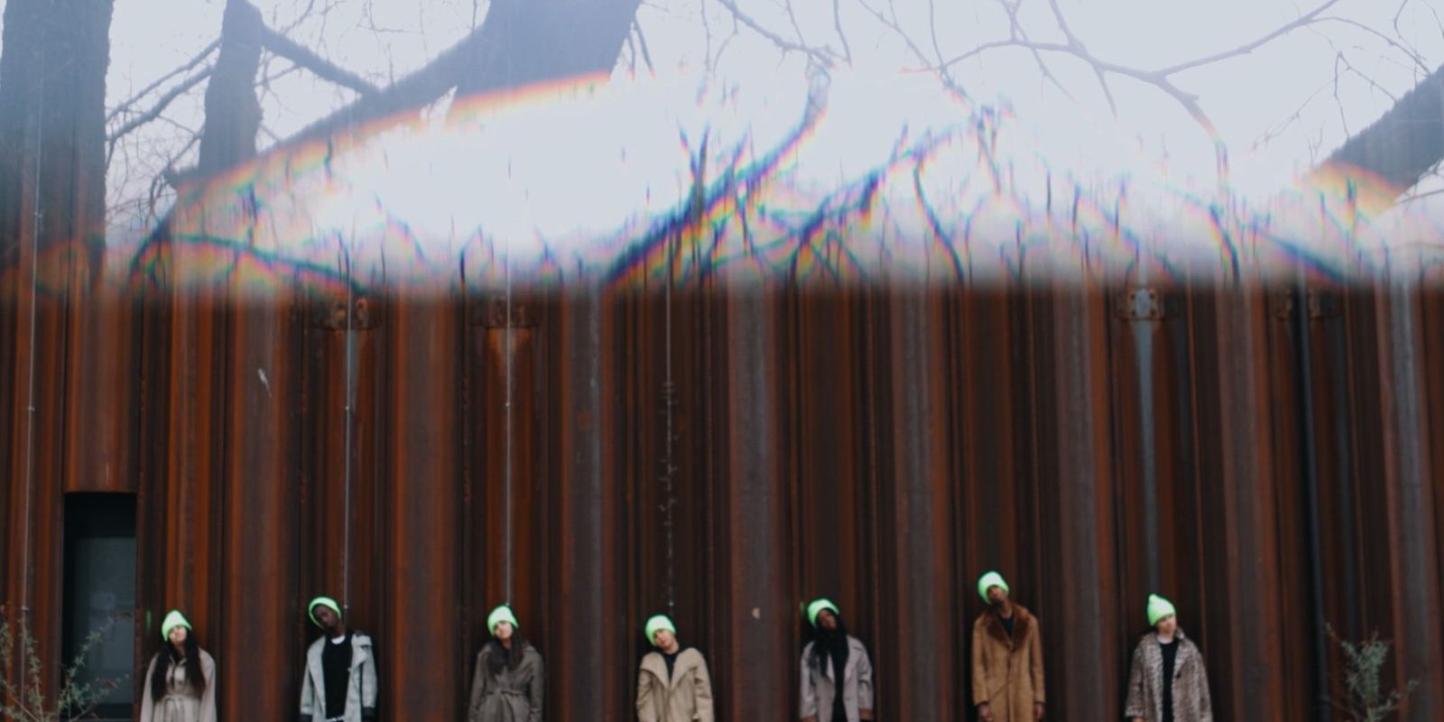 Bottom half of the picture: Seven young people in coats and neon yellow caps stand in front of a rust-coloured building. Each person is standing between a pillar. They all tilt their heads to the left and look forwards. Upper half of the picture: We see bare trees against a white background. This part of the picture is blurred