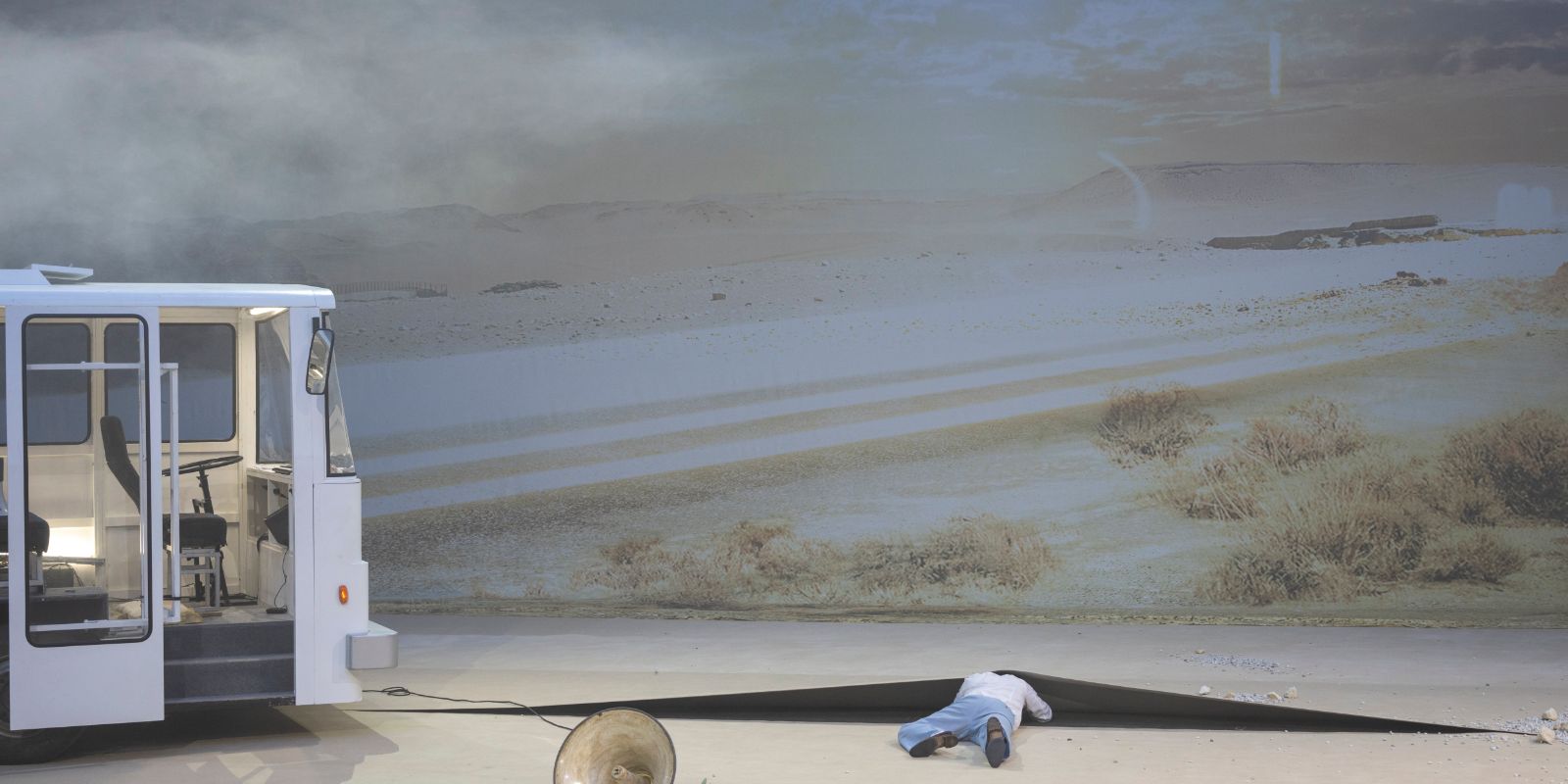 A performer crawls under the light-coloured flooring on a stage with a desert background and a bus on the left.