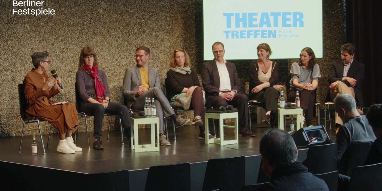 Eight people sit on a stage, one person speaks into a microphone.