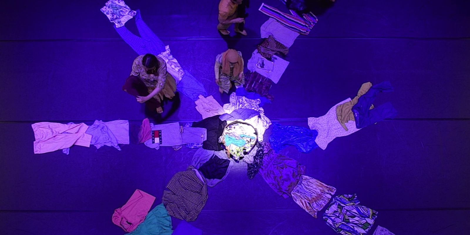 Bird’s eye view of items of clothing arranged in a star shape on the floor. A few people stand between them.