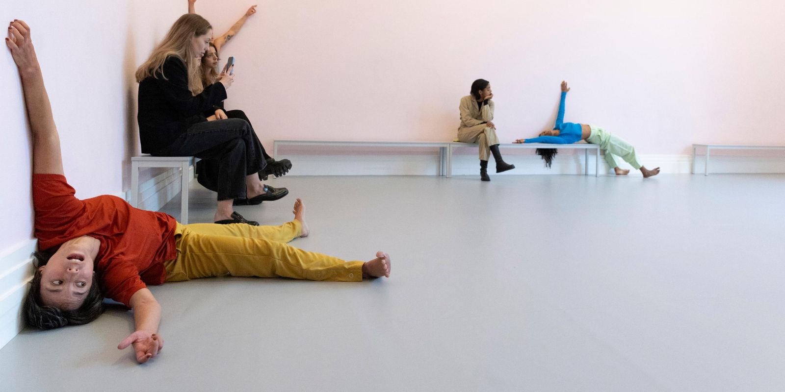 In einem hellrosa gestrichenen Raum findet eine Live-Performance mit dem Titel „study now steady“ statt. Mehrere Tänzer:innen bewegen sich dynamisch durch den Raum und beziehen dabei den Boden, die Wände und die Bänke in ihre Choreografie ein. Das Publikum sitzt auf einfachen Bänken und beobachtet aufmerksam das Geschehen.