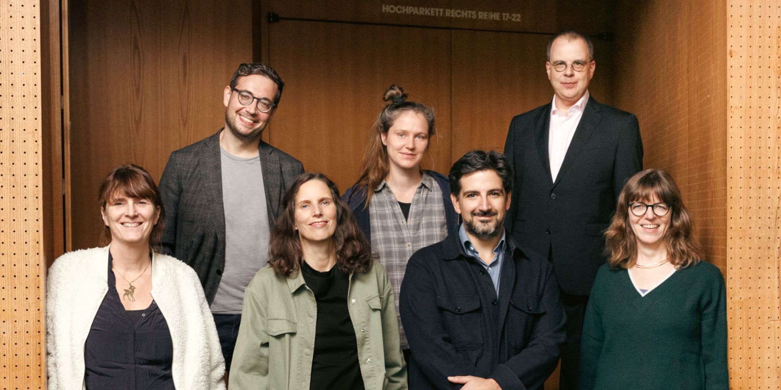 Die sieben Theatertreffen-Jurymitglieder stehen vor einer Saaltür in einem Theater und schauen in die Kamera.