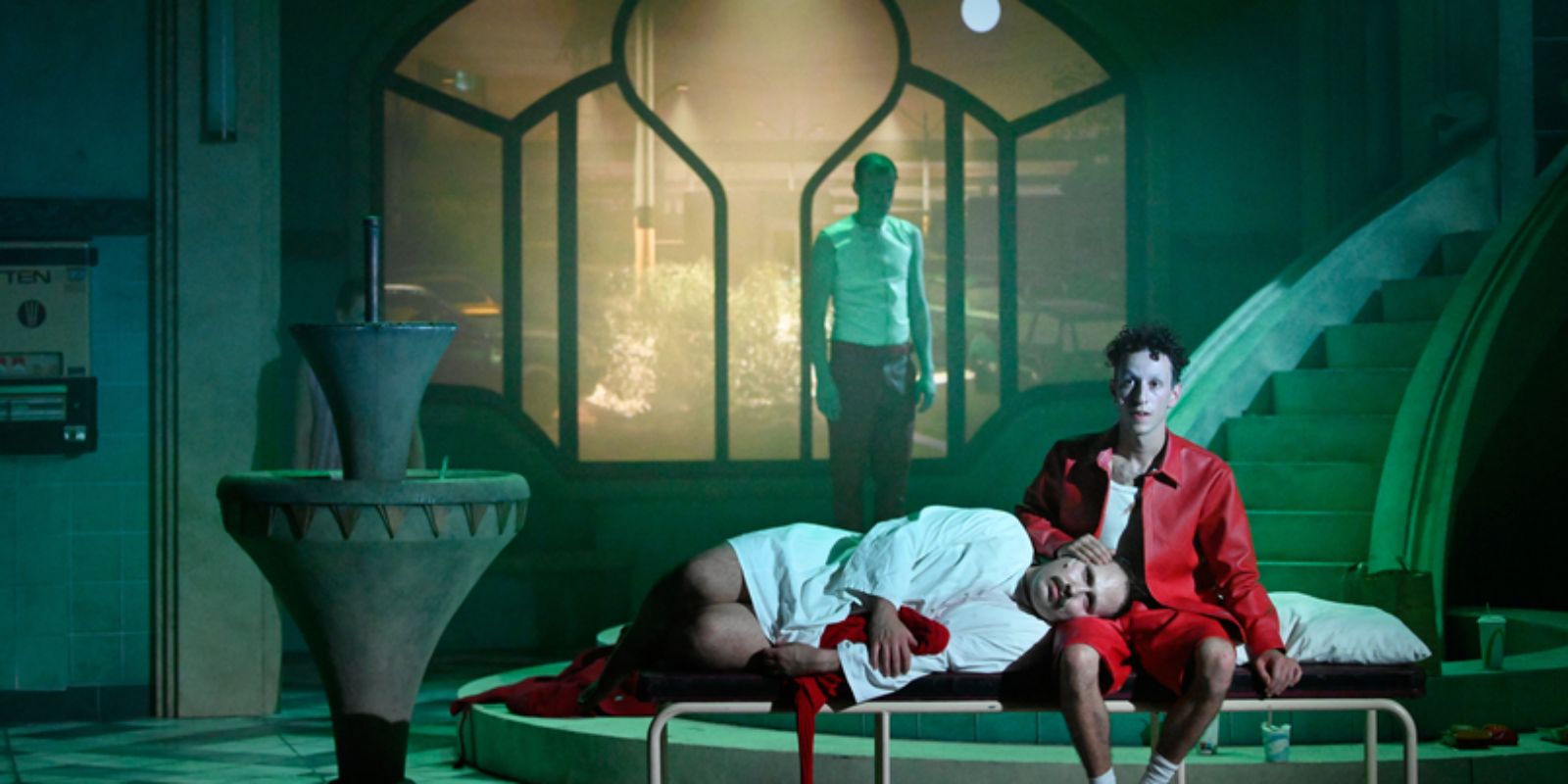 A man in white clothes wearily rests his head on the thigh of a man in a red short suit.