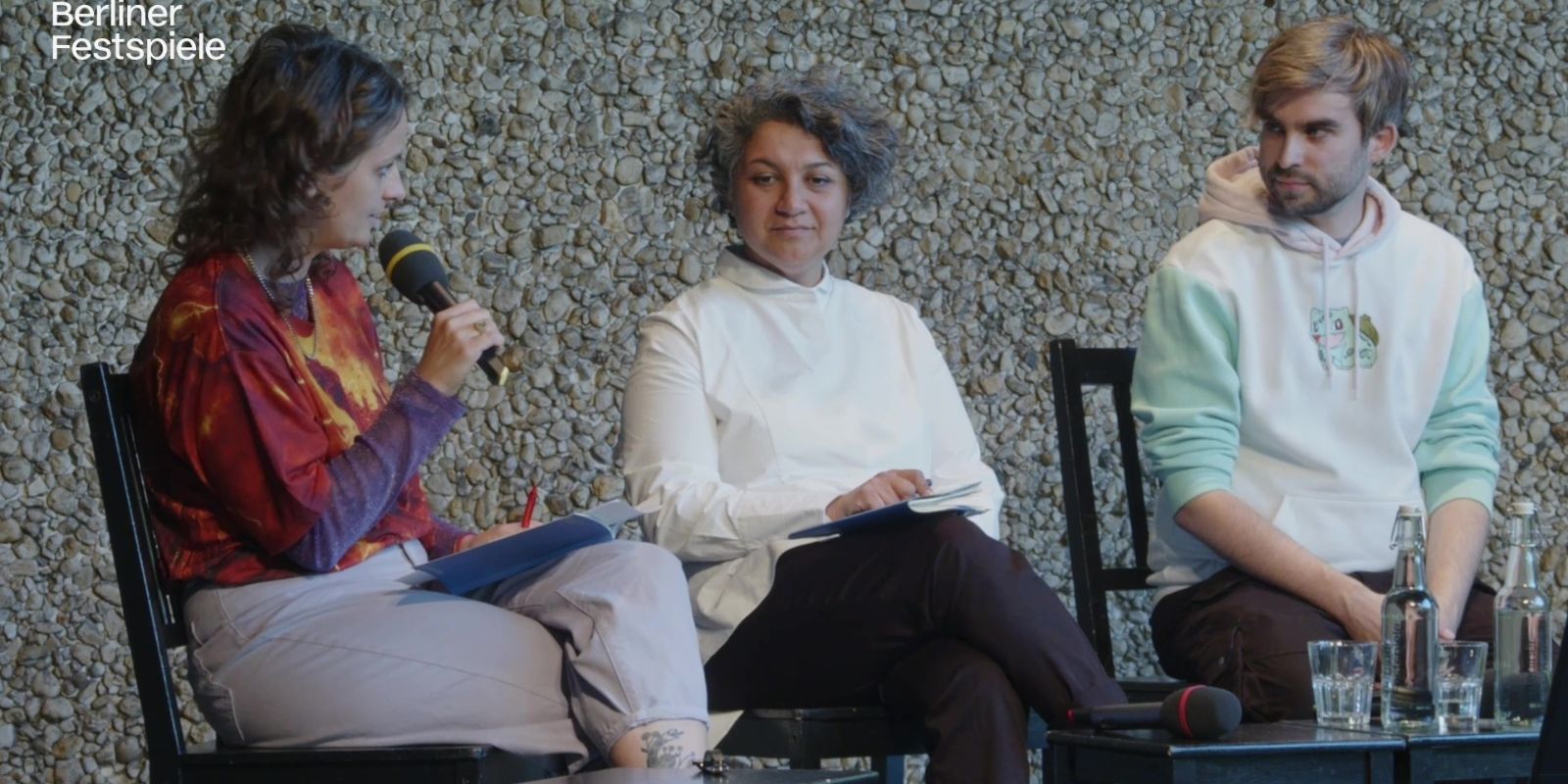 3 Menschen sitzen auf der Bühne, eine Person spricht in ein Mikrofon und die übrigen schauen sie an.