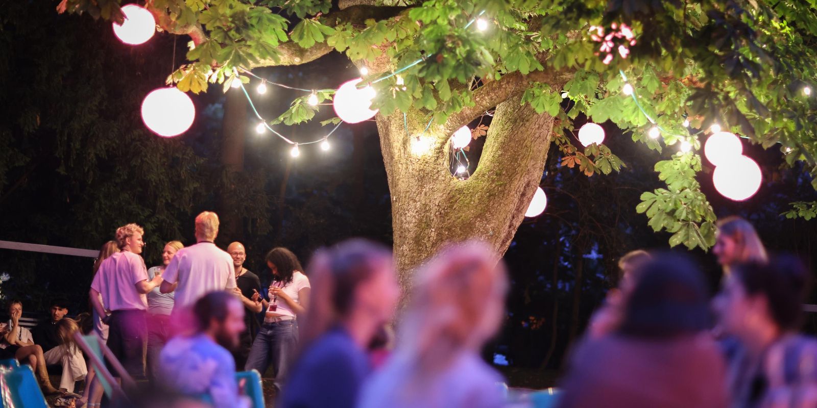 A large tree adorned with colorful lamps takes center stage. In front of it, many young people are standing and sitting in a relaxed atmosphere.