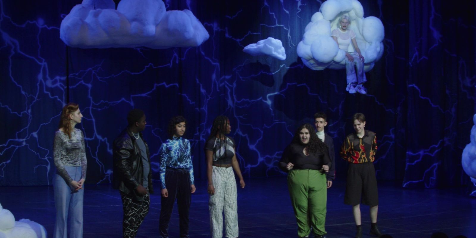 A group of young people stand on a blue-lit stage. Clouds hang from the ceiling.