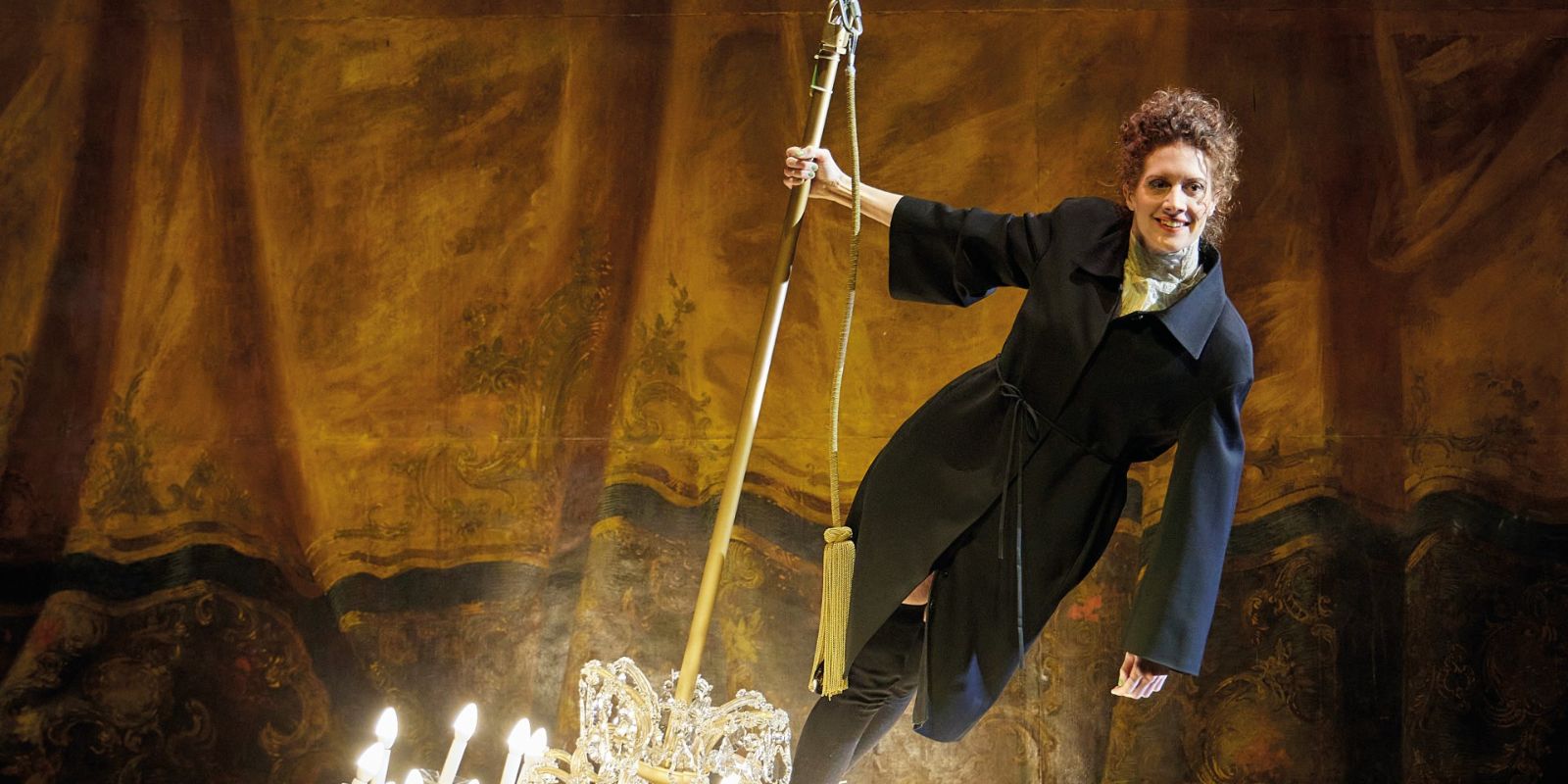 A woman in a blazer‑coat stands smiling on a swinging chandelier