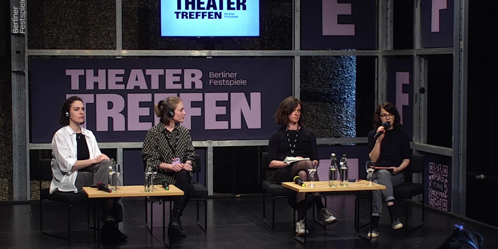 Vier Frauen sitzen an zwei kleinen Tischen, hinter ihnen der Schriftzug Theatertreffen. Die Frau ganz rechts hat ein Mikrofon in der Hand.