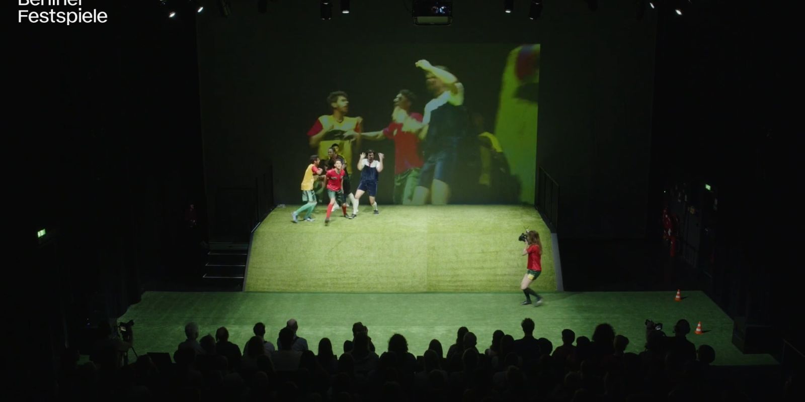 Einige in Fußballtrikots gekleidete Menschen stehen auf einer Erhöhung. Eine Person am Boden filmt. Im Hintergrund wird ein Fußballspiel auf einer Leinwand gezeigt.