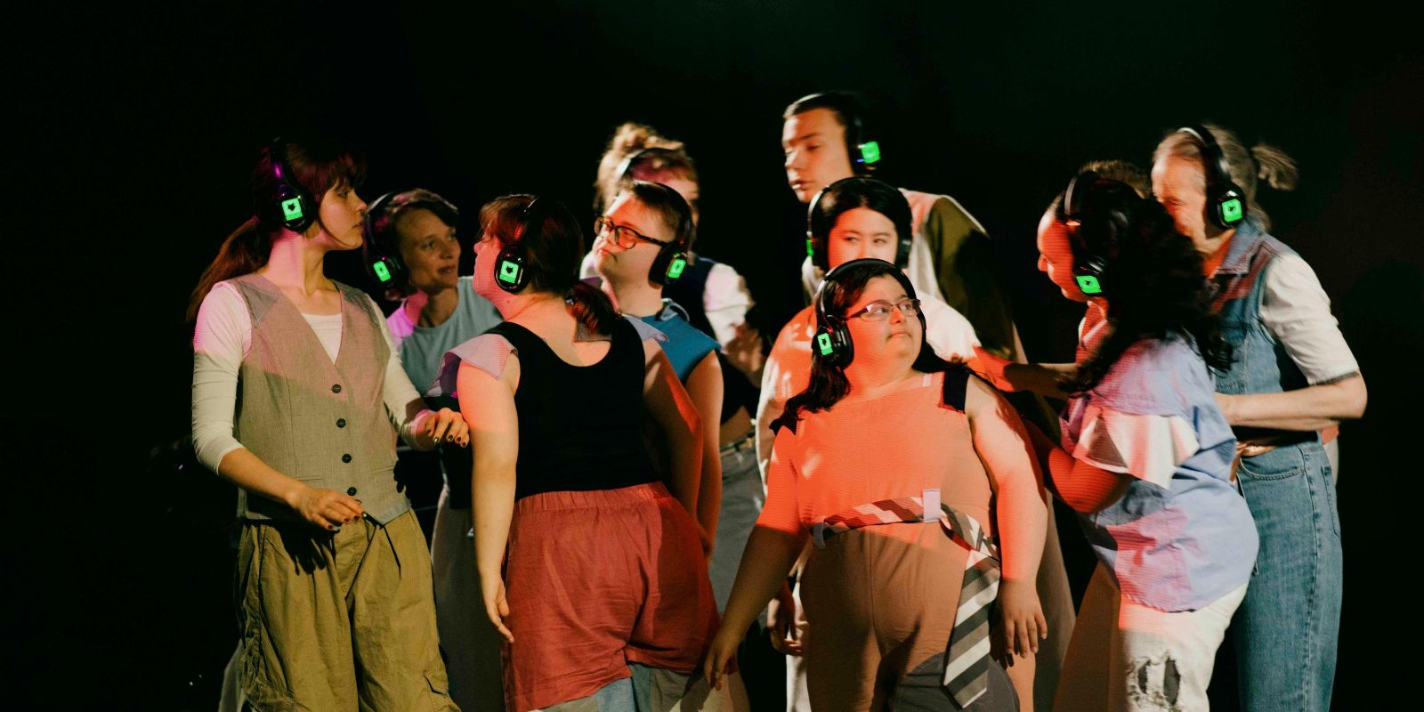 Ein Bühnenfoto mit etwa Zehn bunt angezogenen Personen, die in einem dichten Pulk zusammen stehen. Alle tragen grüne Kopfhorer, wie bei einer Silent-Disko. Die Personen wirken Interresiert und musstern sich gegenseitig. Der Hintergrund ist schwarz.