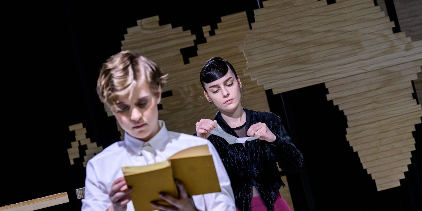 Two young women in old-fashioned costumes are each reading a book. One woman is in the foreground of the picture and out of focus. The second woman is further in the background and is clearly recognisable.