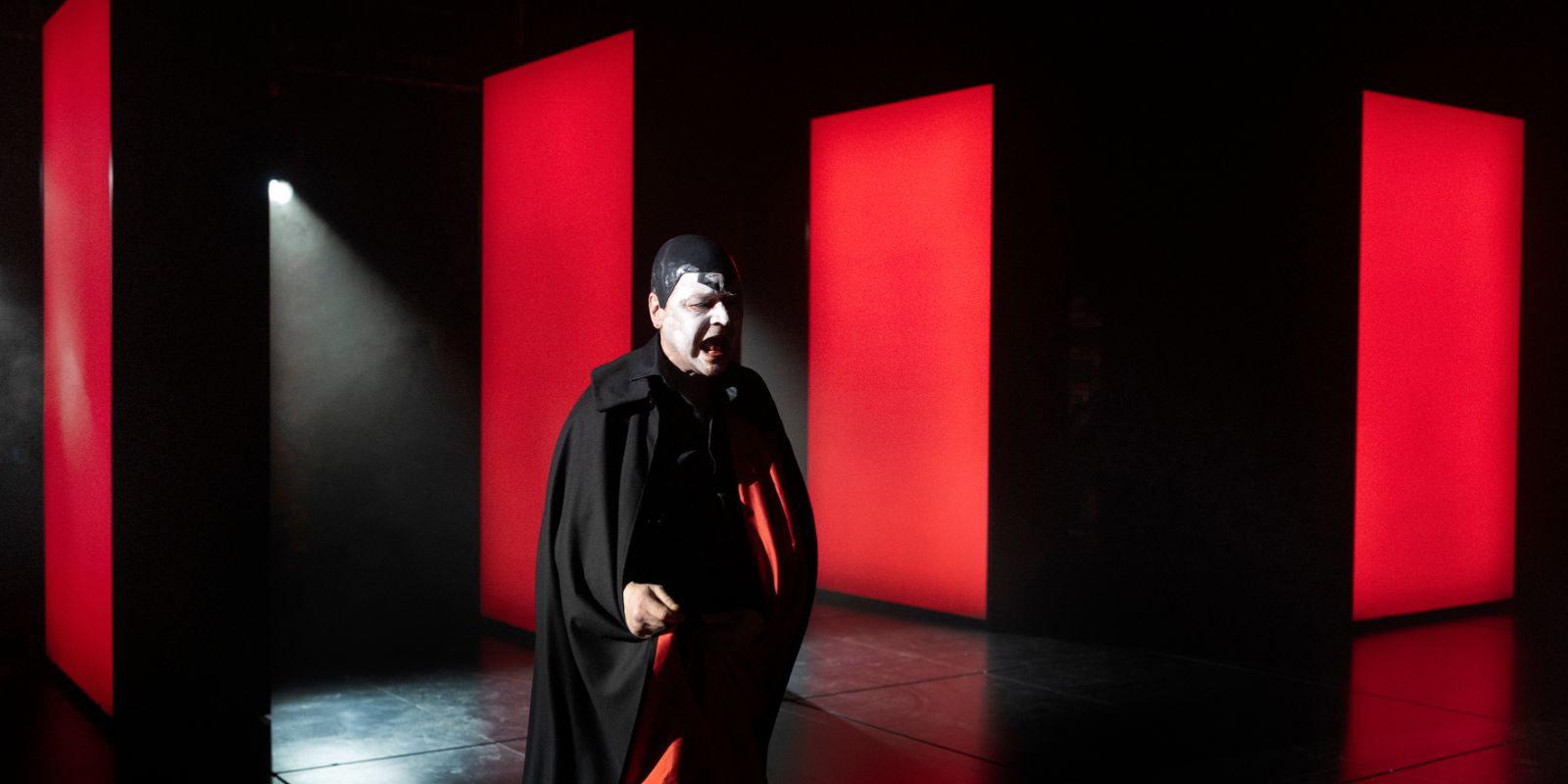 A man with white face paint and a red‑and‑black cloak stands in a red‑and‑black room