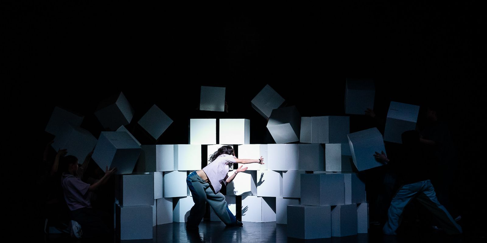 Performers on stage, one in the foreground breaks through a wall made of large white cubes while dancing.