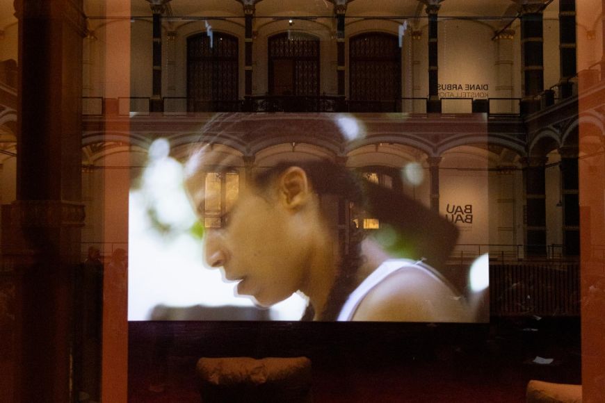 In einem Fenster, mit Blick auf den historischen Lichthof des Gropius Bau, spiegelt sich ein Bildschirm. Zu sehen ist eine Szene aus der Arbeit Arbeit „A Plot, A Scandal“ der der Künstlerin Ligia Lewis. Das angeschnittene Profil von Ligia Lewis ist schemenhaft zu erkennen.