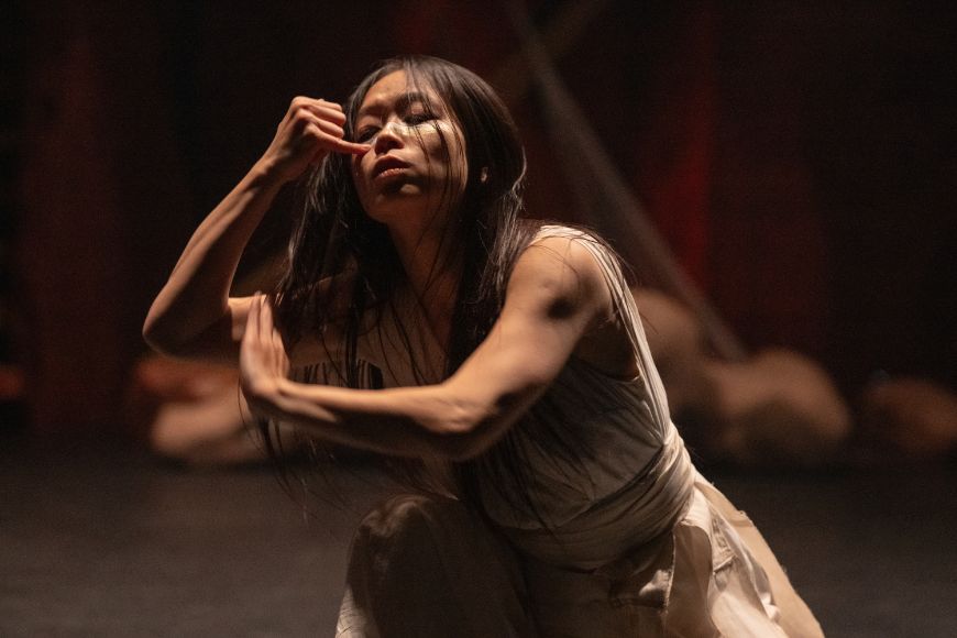 A dancer in a simple dress concentrates on painting her face with colour.