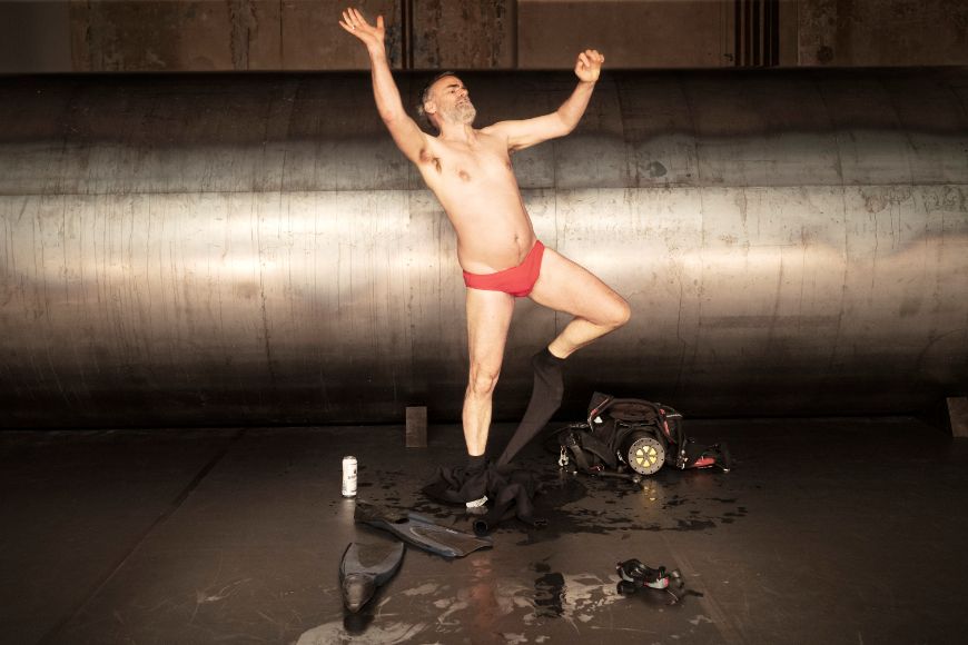 Eine schauspielende Person strampelt sich stehend aus einer Hose und trägt darunter einen Slip.