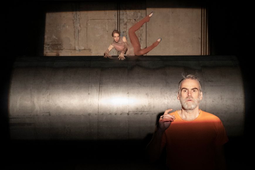 Eine Person posiert sitzend und mit den Beinen in der Luft. Im Vordergrund blickt eine Person erstaunt.