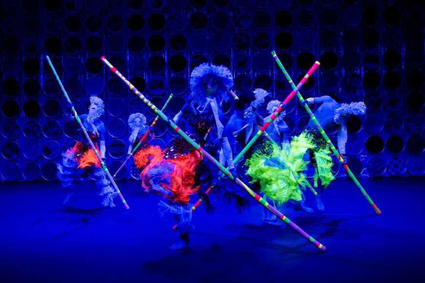 The dancers' frilly costumes and long poles glow in bright neon colours.