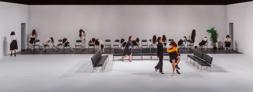 In einem weiten Raum stehen zahlreiche Stühle. Auf manchen sitzen lebensgroße Puppen. Zwei Menschen umarmen sich.