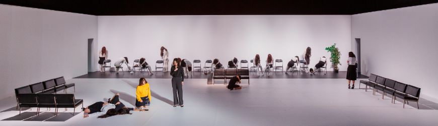 Leicht düsteres Bühnenbild, das einer Wartehalle nachempfunden ist. Zwei Personen befinden sich neben zwei am Boden liegenden Tänzerinnen.