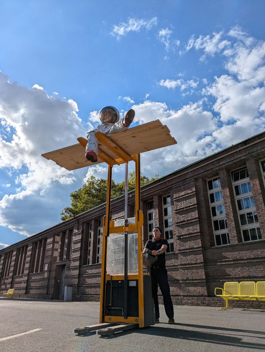 Eine Schauspielerin im Austronautinnen-Kostüm sitzt erhöht auf einem Gabelstapler.