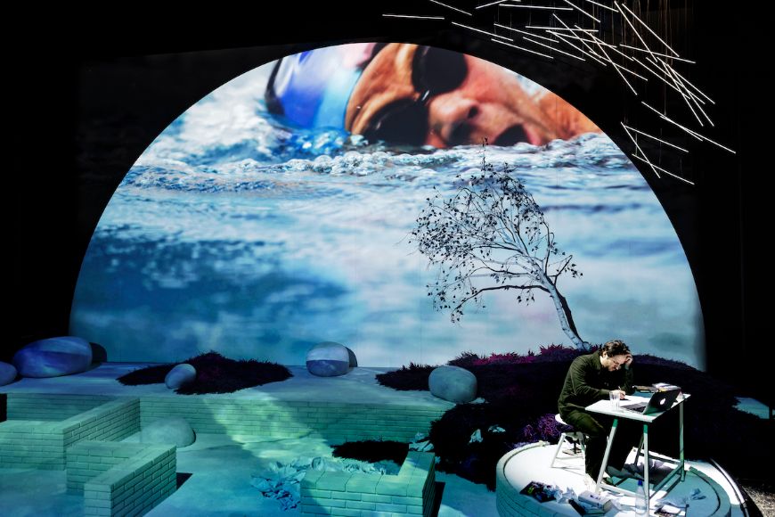 Auf der Bühne eine Person am Schreibtisch, dahinter die Projektion einer Schwimmerin