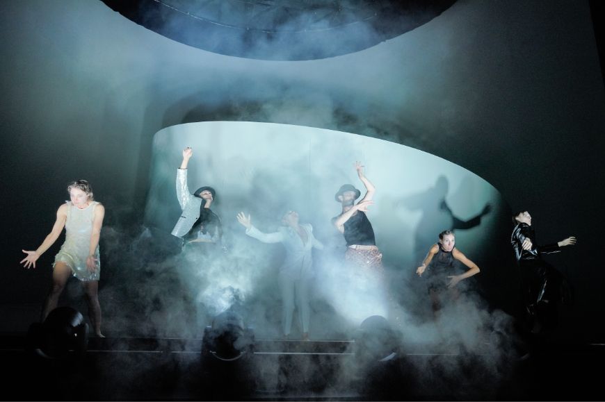 Six people dance expressively in stage fog in front of a white spiral stage set.