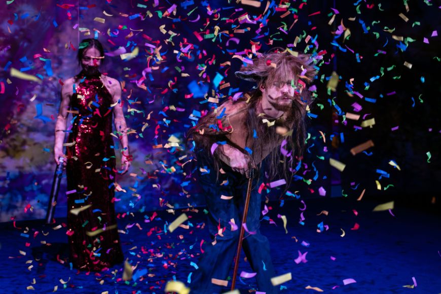 A man leans on a stick on a stage where confetti is raining down. Behind him is a woman in an evening dress holding some kind of stick.