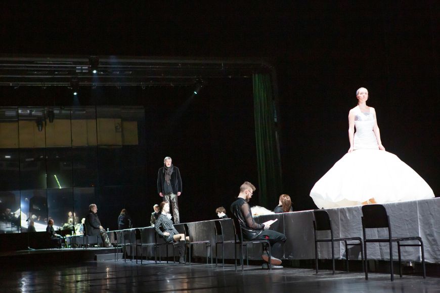 Eine Frau in einem weißen ausladenden Kleid schreitet über einen langen Tisch, an dem rechts und links Personen sitzen.