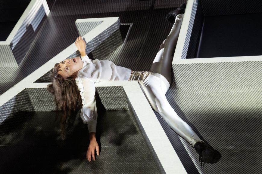 A woman with long hair lies with twisted limbs in a stage set with several shelf-like elements.