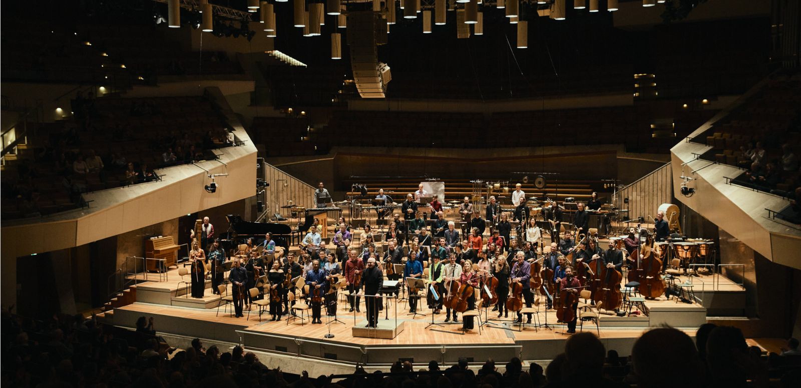 An orchestra stands on the brightly lit stage in a darkened concert hall.