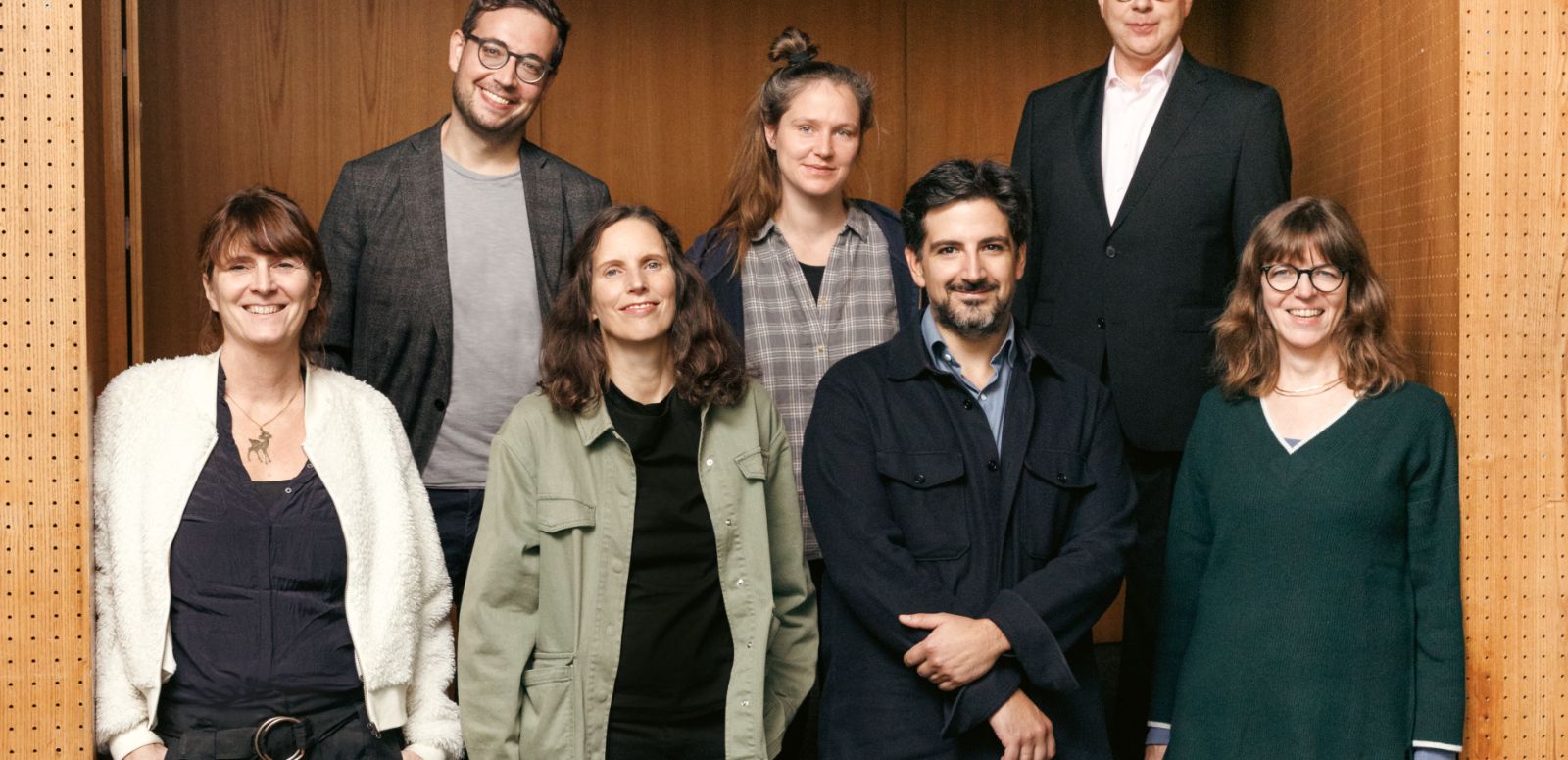 The seven Theatertreffen jury members stand in front of a door of a theatre auditorium and look into the camera.